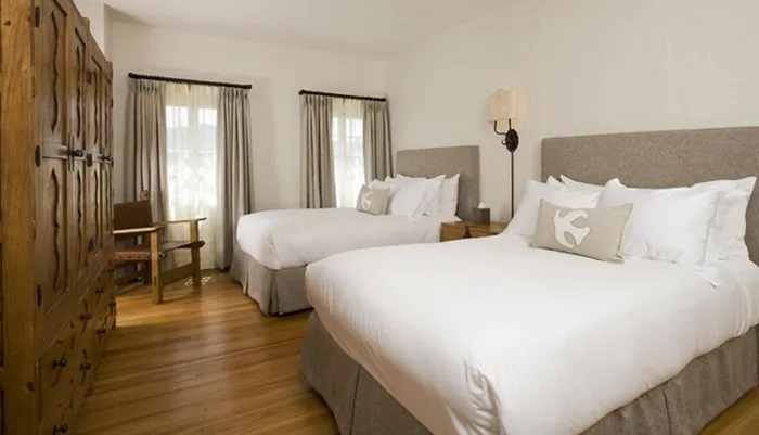 This image shows a neatly arranged hotel room with two beds crisp white bedding wooden floors and a rustic wooden wardrobe