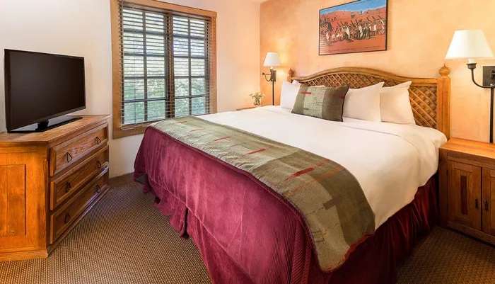 This is an image of a well-appointed bedroom with a large bed covered in white linens and a red bedspread wooden furniture a flat-screen TV and a painting above the headboard