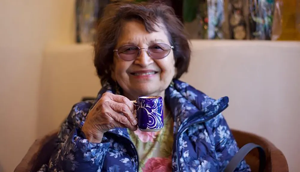 An elderly woman with glasses is smiling warmly at the camera while holding a small decorative blue cup