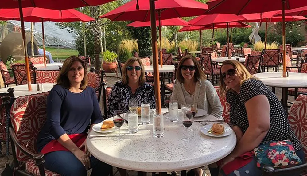 Four women are smiling at the camera while seated at an outdoor patio table with red umbrellas enjoying what appears to be a pleasant meal together