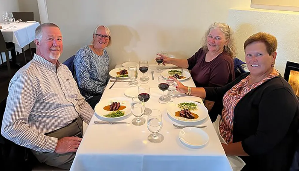 Four people are smiling at the camera while seated around a dining table with plates of food and glasses of wine in a restaurant setting