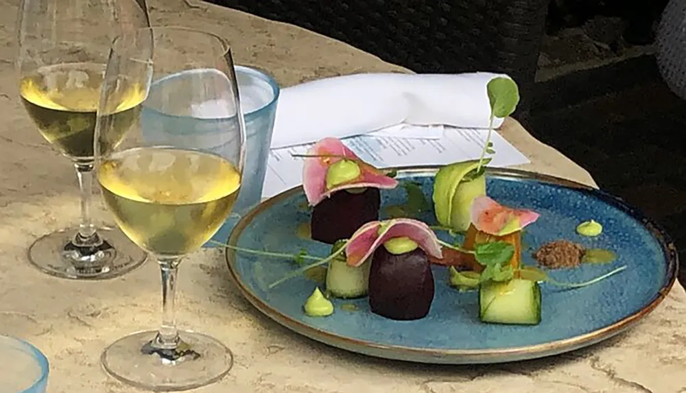 The image shows an elegantly plated dish with colorful garnishes accompanied by two glasses of white wine suggesting a fine dining experience