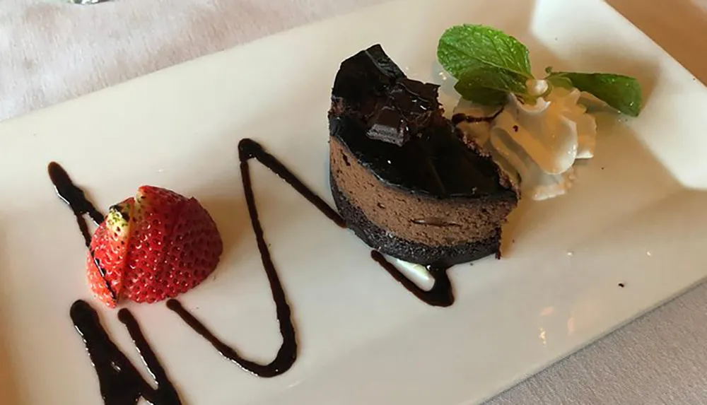A slice of chocolate cake with a glossy chocolate topping is elegantly presented on a plate with a fresh strawberry and a decorative drizzle of chocolate sauce