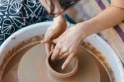 A person is shaping clay on a pottery wheel, their hands and the wheel covered in wet clay.