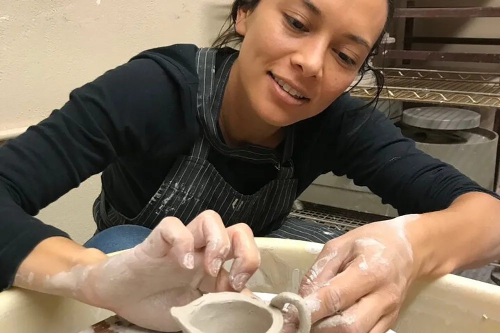 A person is happily engaged in shaping a clay pot on a pottery wheel with their hands covered in clay
