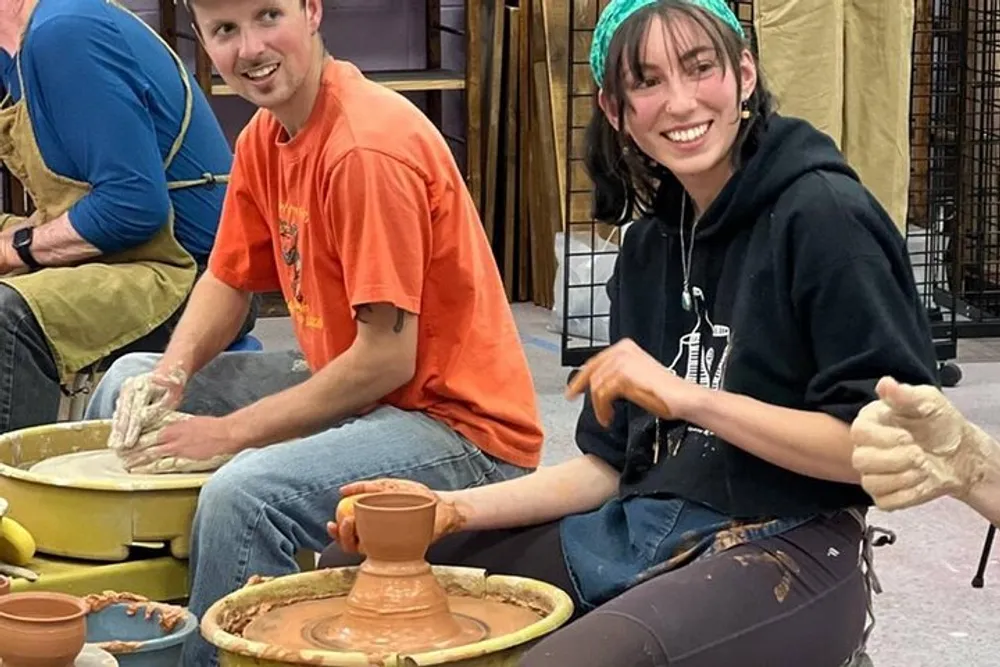 Two people are smiling at the camera while engaged in pottery making at a crafting workshop with one showing a completed clay pot on the wheel