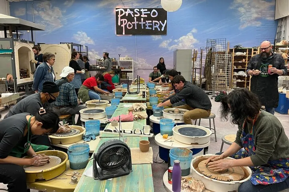 A group of people is engaged in pottery making at a workshop with a sign that reads PASEO POTTERY