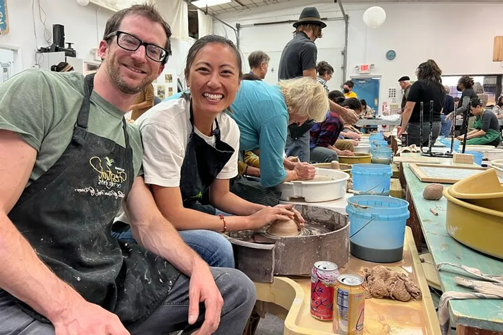 A man and a woman are smiling for the camera while seated at a pottery workshop with several other participants focused on their craft in the background