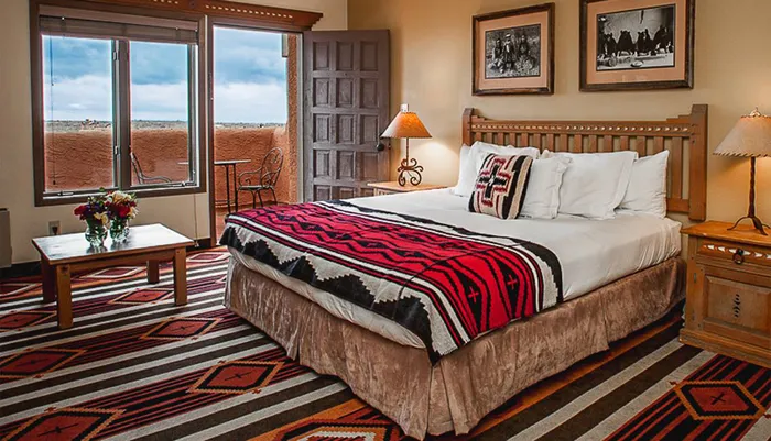 The image shows a warmly decorated bedroom with a large bed adorned with a colorful geometric-patterned blanket complementing the Southwestern-style rug on the floor and with a scenic view through a window