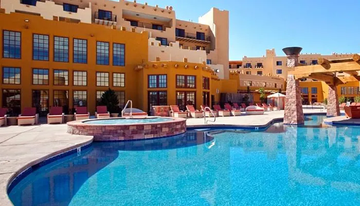 The image shows a bright and sunny pool area with loungers and a Southwestern-style hotel building in the background