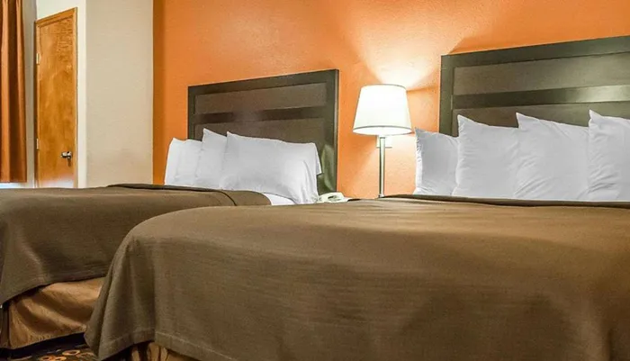 The image shows a neatly arranged hotel room with two double beds a bedside lamp and a warm color scheme