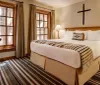 The image shows a warmly lit cozy bedroom with a large bed earthy tones striped carpet wooden furniture and a cross hanging on the wall