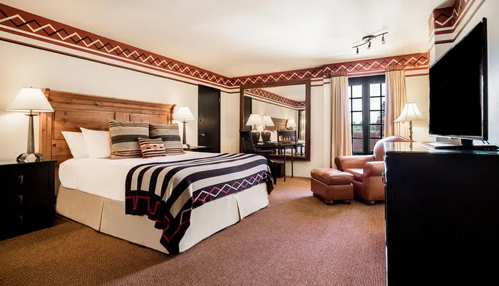 The image shows a well-appointed hotel room with Southwestern or Native American design accents featuring a large bed with patterned blankets comfortable seating and a flat-screen TV