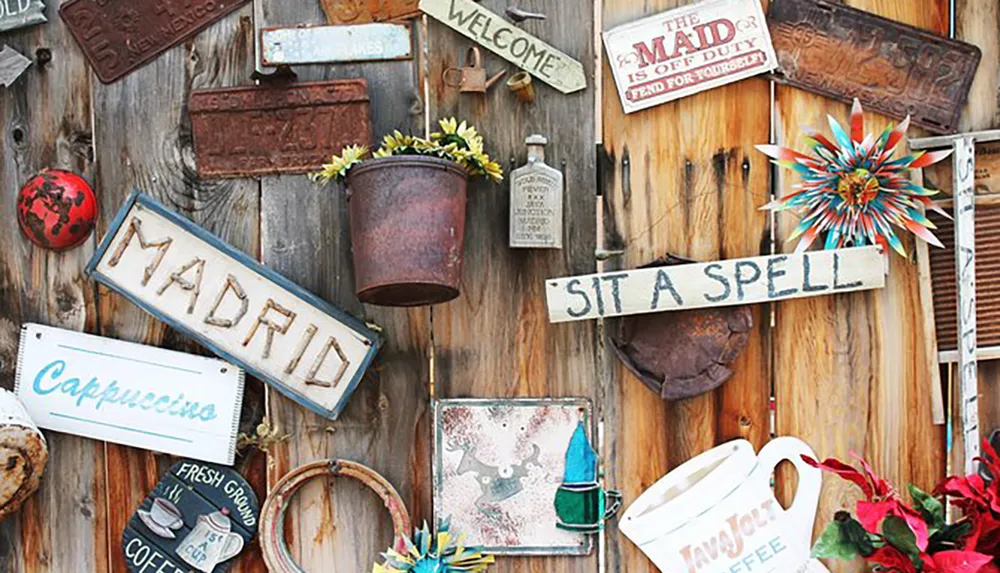 The image shows a rustic wooden wall adorned with a variety of eclectic and vintage signs decorative plates a colorful metal star and pots with flowers creating a charming and whimsical display