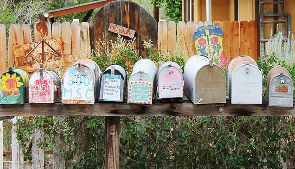 A row of colorful and uniquely decorated mailboxes lines a wooden shelf against a rustic fence backdrop