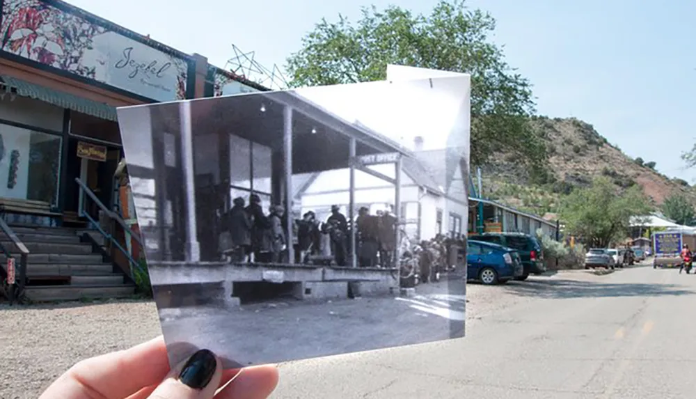 A persons hand is holding up a black and white photo of an old post office blending the past scene with the current street view in color