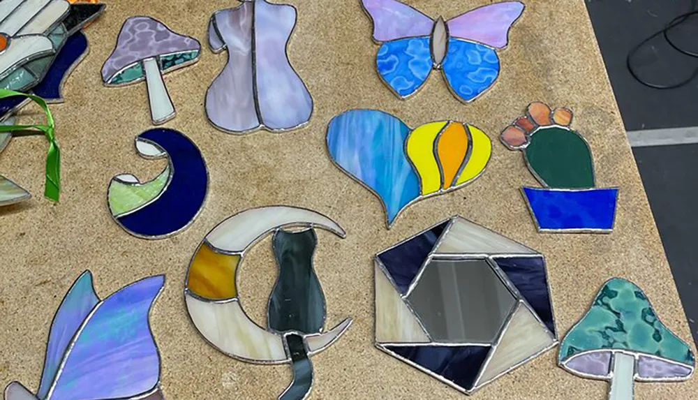 The image shows a collection of colorful stained glass pieces in various shapes including plants and abstract patterns laid out on a corkboard-like surface
