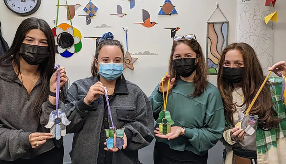 Four individuals wearing masks are proudly holding up colorful stained glass creations against a backdrop decorated with artistic elements