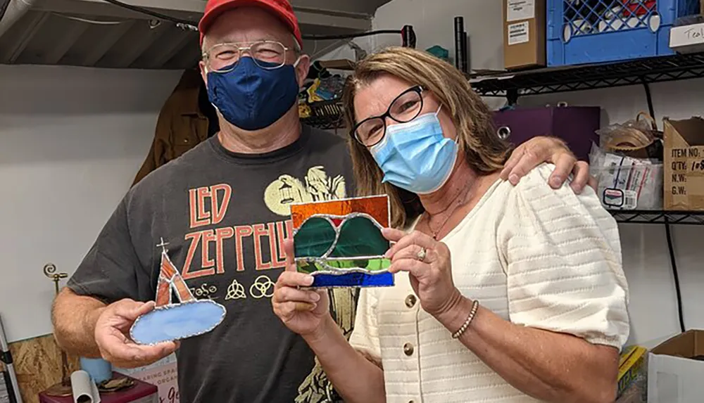 Two smiling people wearing face masks are holding colorful stained glass artworks in a room that appears to be a workshop or storage area