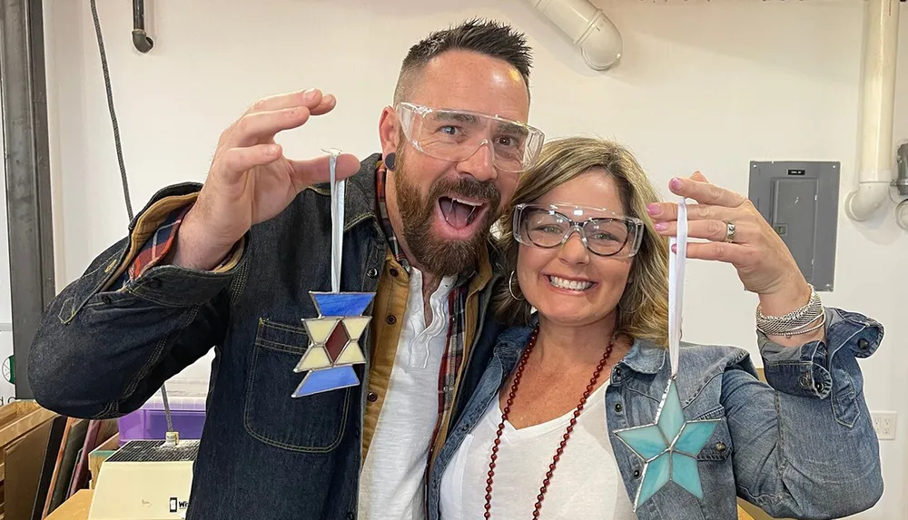 A man and a woman are smiling and posing with star-shaped glass art pieces wearing protective eyewear likely inside a workshop