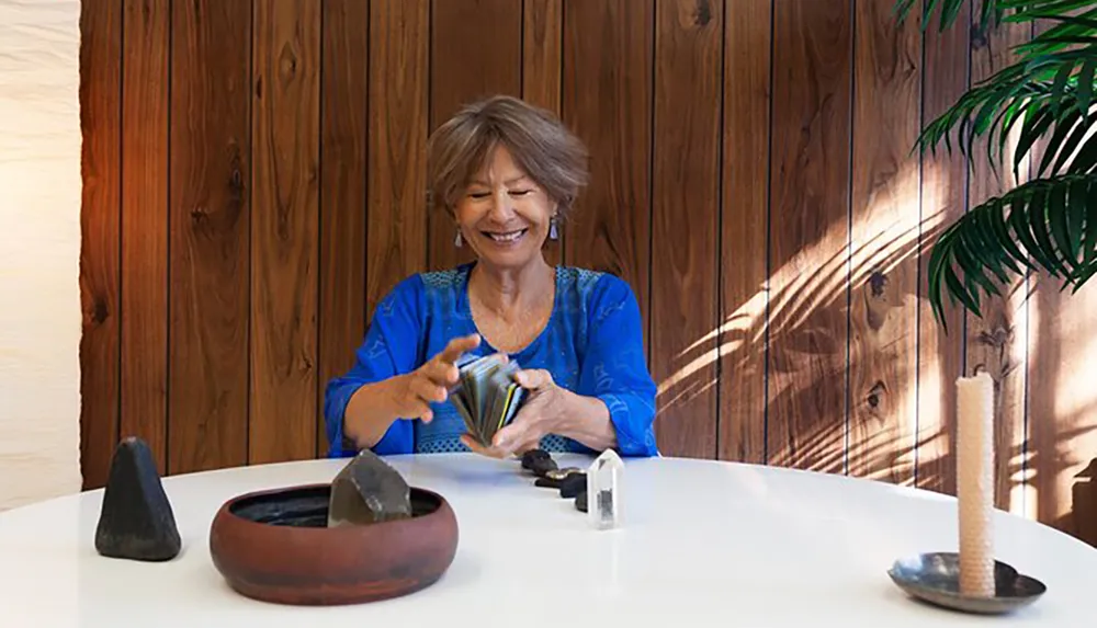 A smiling woman is handling a deck of cards at a table with a rustic wooden backdrop and some decorative items creating a serene setting