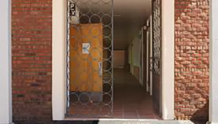 The image shows a rustic yellow door with iron grilles, flanked by two exposed brick walls, leading into a corridor with arches.