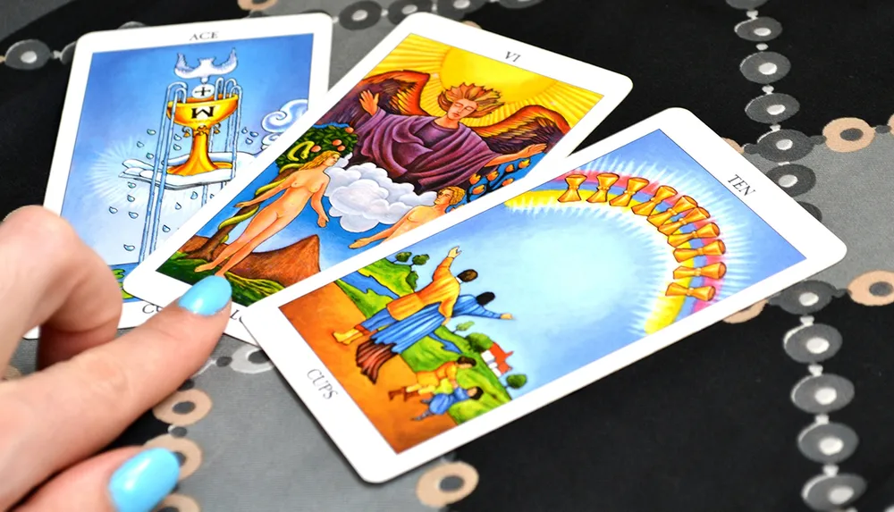 A person is displaying three tarot cards on a patterned table with the Ace of Cups the Lovers and the Ten of Cups revealed