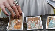 A hand adorned with rings touches a tarot card laid on a surface among other scattered cards.