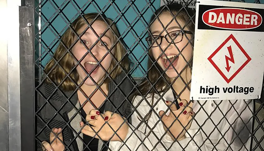 Two people are playfully posing behind a chain-link fence with a DANGER high voltage sign