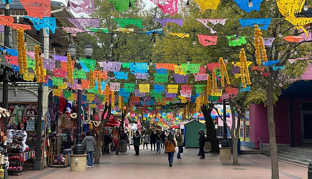 The image shows a vibrant outdoor market adorned with colorful papel picado banners and marigold garlands with people browsing the various stalls
