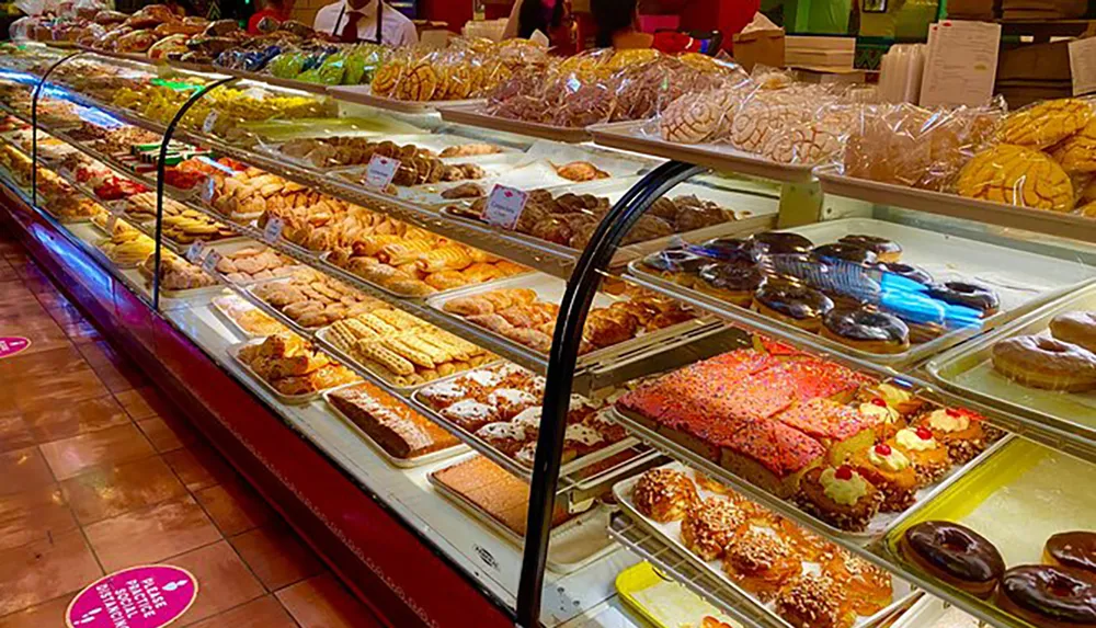 The image shows a bakery display case filled with an assortment of colorful and freshly baked pastries and cookies with shoppers in the background