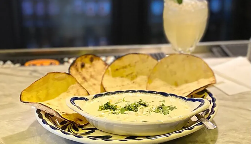 A bowl of creamy dip garnished with herbs is served with large toasted chips accompanied by a refreshing drink in the background