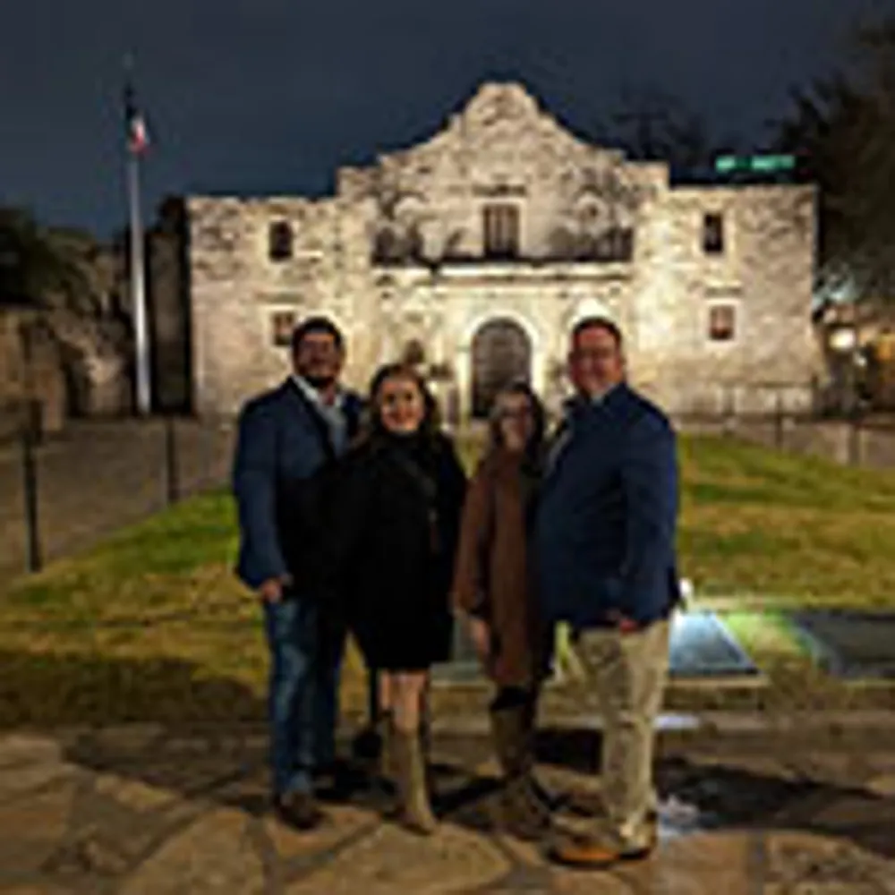 A group of four people are posing in front of the Alamo at night