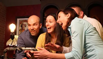 Four people are excitedly gathered around a mysterious, vintage-looking device, with expressions ranging from amazement to amusement.