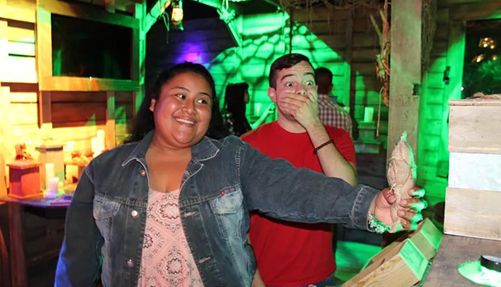 A woman is smiling and holding an object while a man beside her covers his mouth with his hand appearing surprised in a room lit with green and purple lights