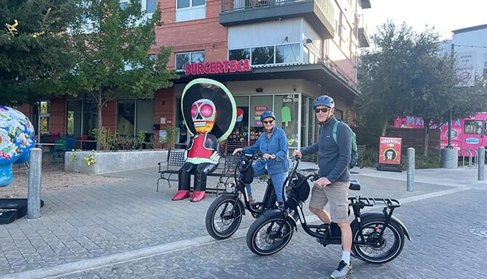 Two people wearing helmets are standing with their bicycles in front of a colorful art installation and a restaurant named BURGERTECA