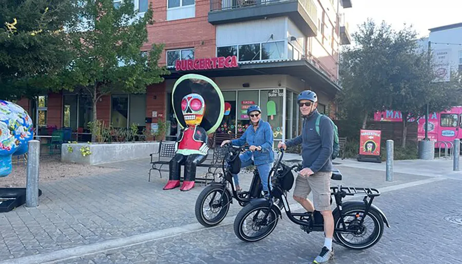 Two people wearing helmets are standing with their bicycles in front of a colorful art installation and a restaurant named BURGERTECA.