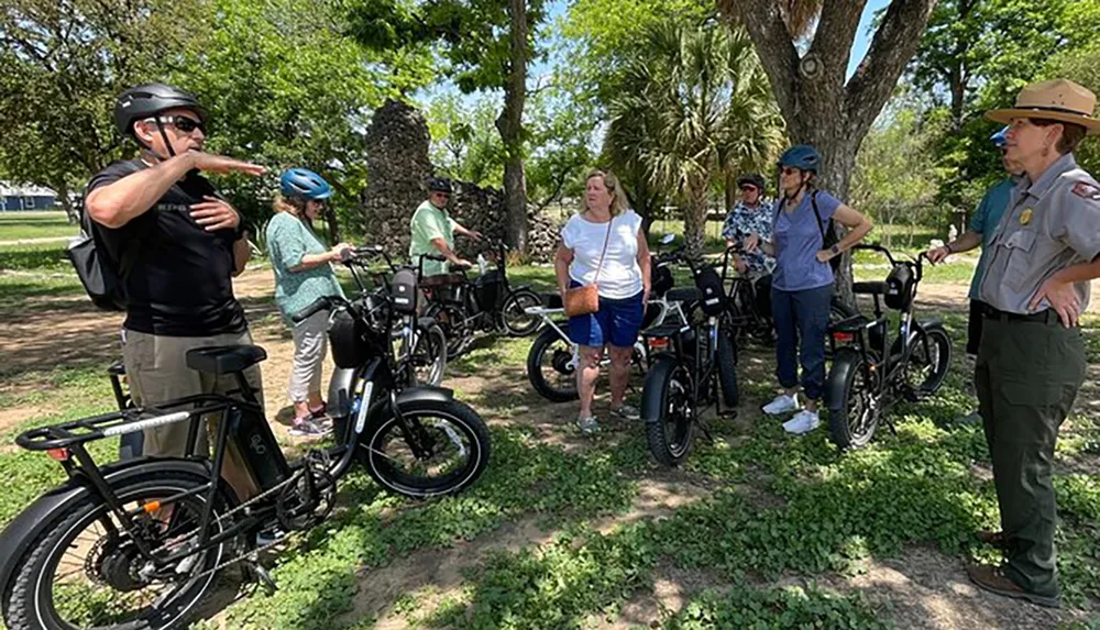A group of people wearing helmets and casual clothing stand around electric bicycles listening to someone gesturing and speaking outdoors indicating a possible guided tour or outdoor activity in a park-like setting