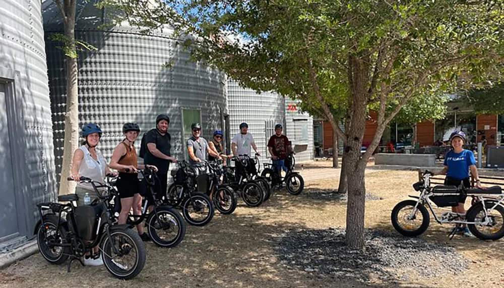 A group of people wearing helmets are standing with their electric bikes in a sunny outdoor setting possibly preparing for or taking a break from a group ride