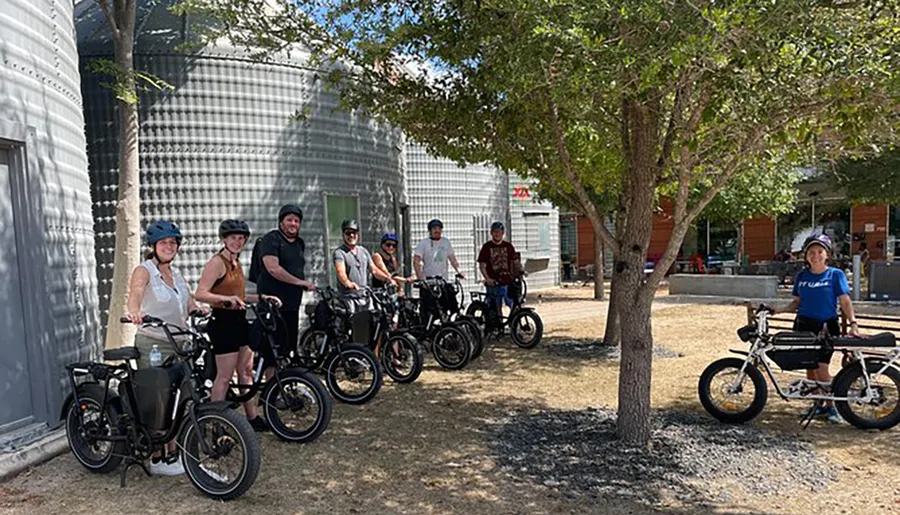 A group of people wearing helmets are standing with their electric bikes in a sunny, outdoor setting, possibly preparing for or taking a break from a group ride.