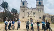 A group of people on electric bikes are posing for a photo in front of an old church with two bell towers.