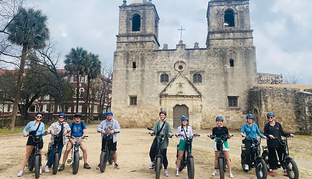 A group of people on electric bikes are posing for a photo in front of an old church with two bell towers