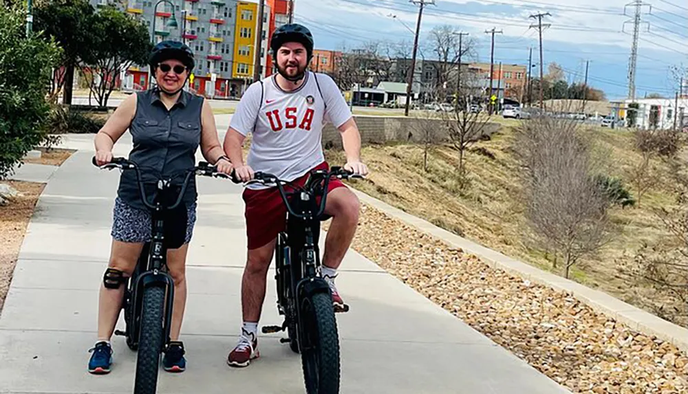 Two people are enjoying a bike ride together on a paved path with urban architecture in the background