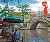 This vibrant image depicts people enjoying outdoor dining by a canal with colorful umbrellas and a bridge in the background beneath a large abstract sculpture