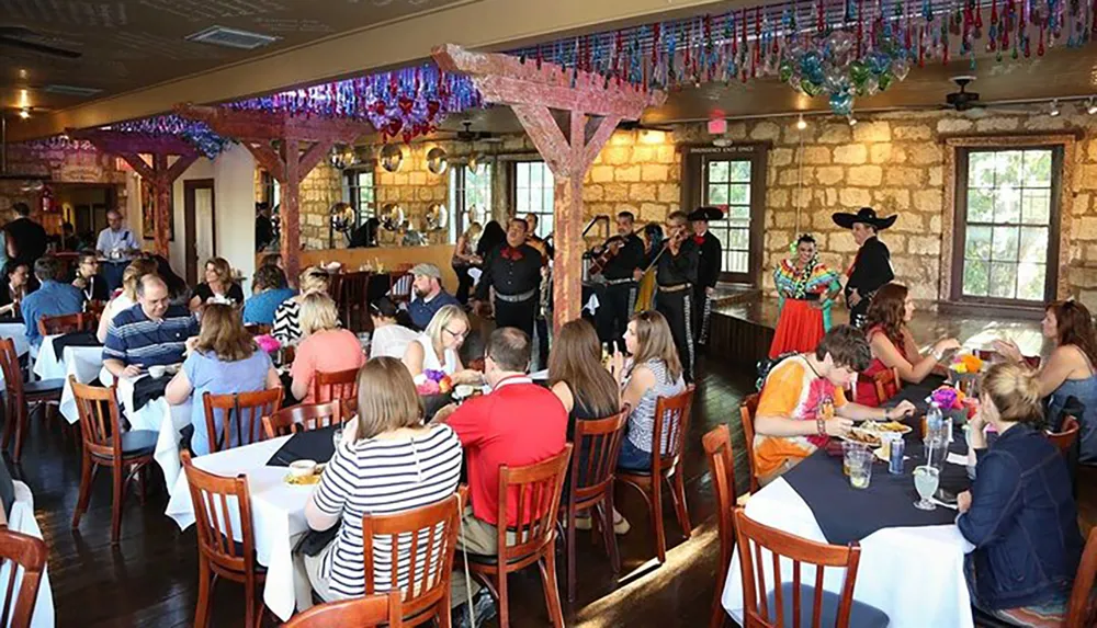 The image shows a lively scene inside a restaurant where patrons are dining and being entertained by musicians and dancers dressed in traditional folk attire