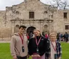 Three smiling visitors are posing for a photo in front of the historic Alamo Mission in San Antonio Texas