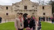 Three smiling visitors are posing for a photo in front of the historic Alamo Mission in San Antonio, Texas.