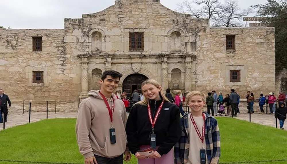 Three smiling visitors are posing for a photo in front of the historic Alamo Mission in San Antonio Texas