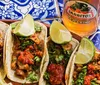 The image shows three flavorful tacos garnished with lime on a colorful patterned surface accompanied by a bottle of Jarritos Tamarind soda