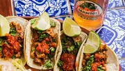 The image shows three flavorful tacos garnished with lime on a colorful patterned surface, accompanied by a bottle of Jarritos Tamarind soda.
